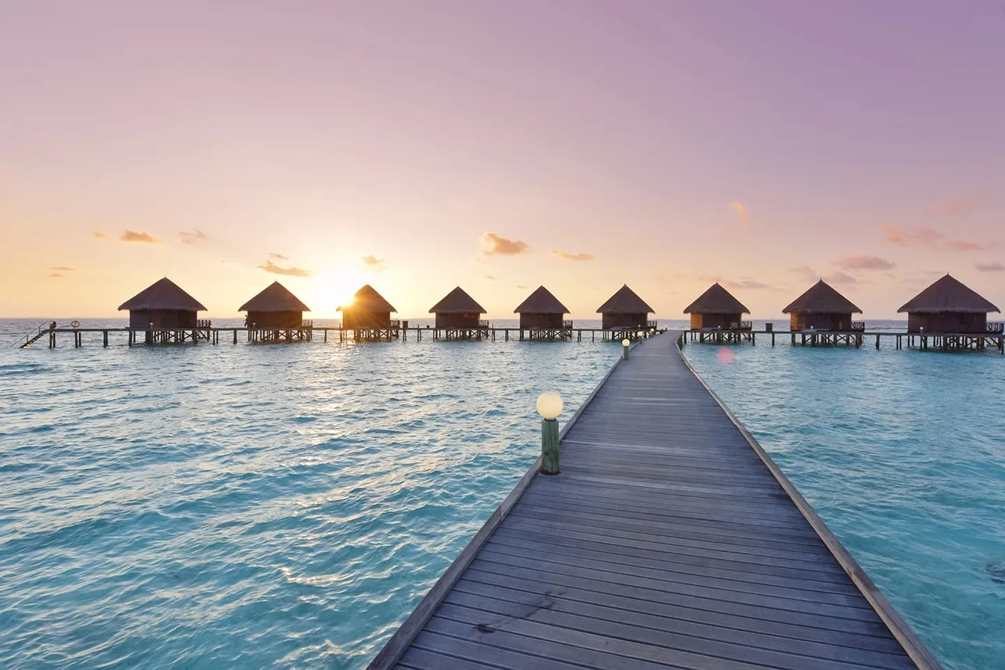 The jetty leading to the water bungalows at sunrise, the sky pink and the sun between the roofs.