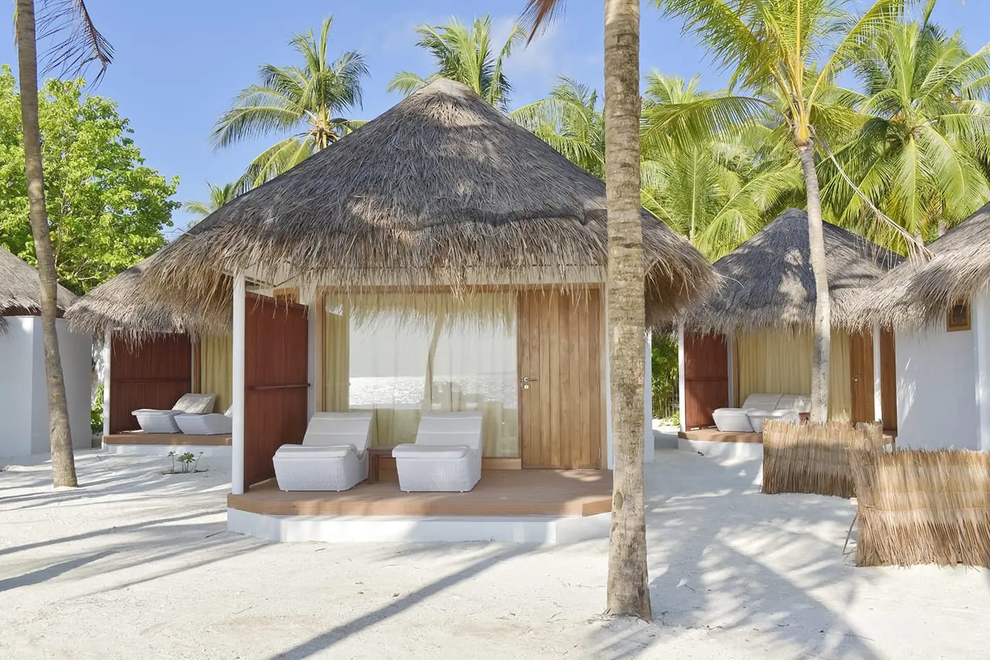 A thatched beach bungalow with its wooden deck and 2 white loungers among the palms.