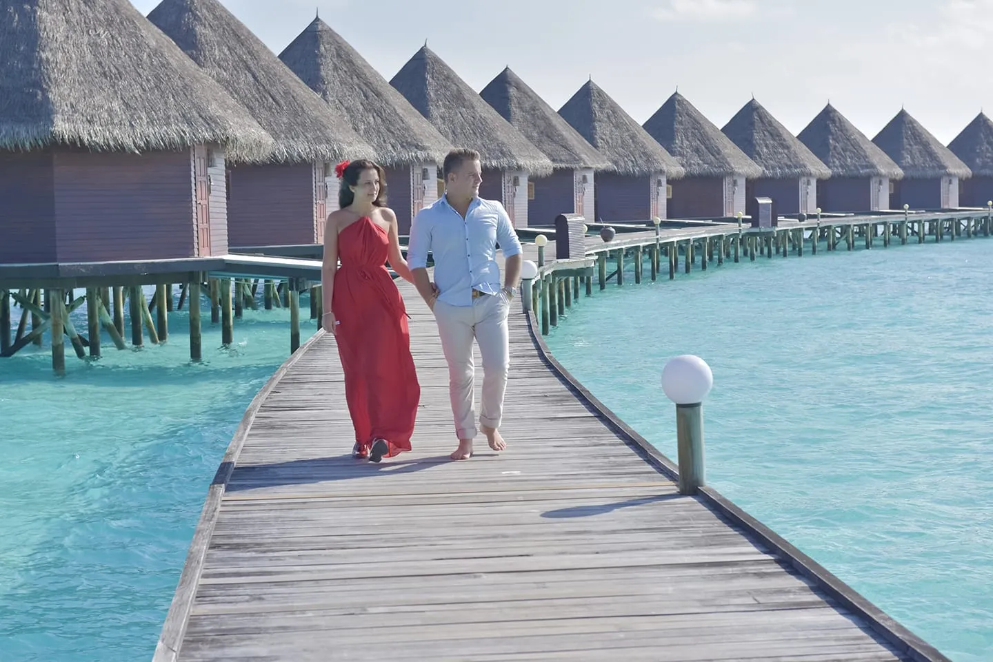 A couple walk along the wooden jetty past the water bungalows, she in a red dress.