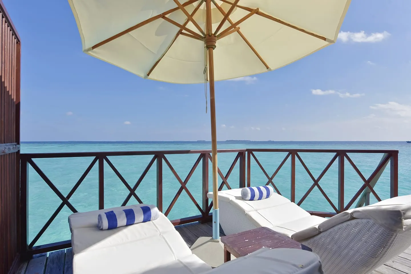 A water bungalow deck: 2 loungers with striped cushions under the parasol, the lagoon beyond the railing.
