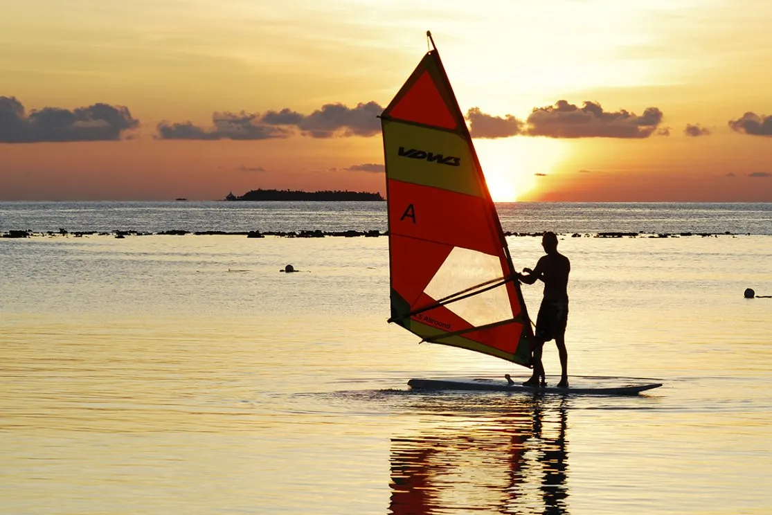 A windsurfer standing in the shallows at sunset, the red sail against the light.