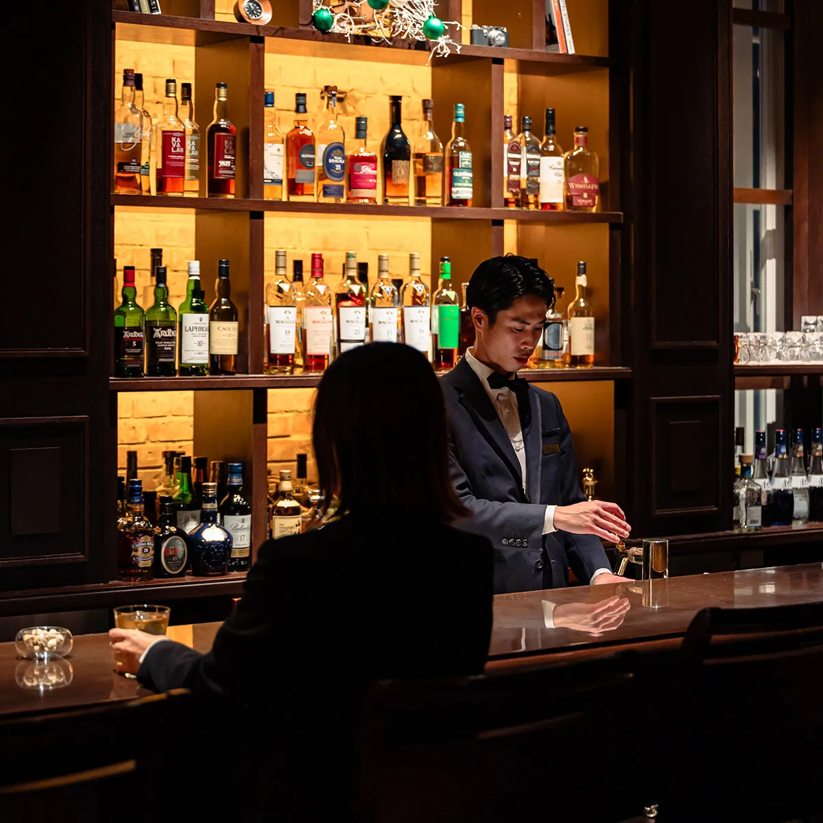 The counter at Bar Oak: a bartender in a jacket mixes a cocktail in front of the whisky shelves, with a guest seated at the bar, seen from behind.