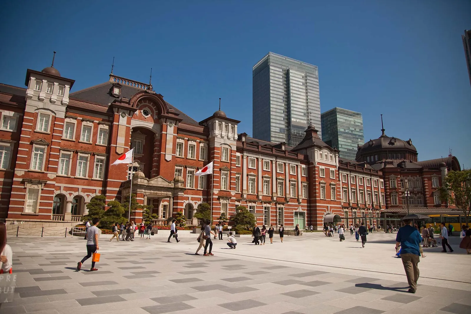The station’s red-brick façade seen from the Marunouchi square, with 2 flags, a blue sky and a few passers-by.