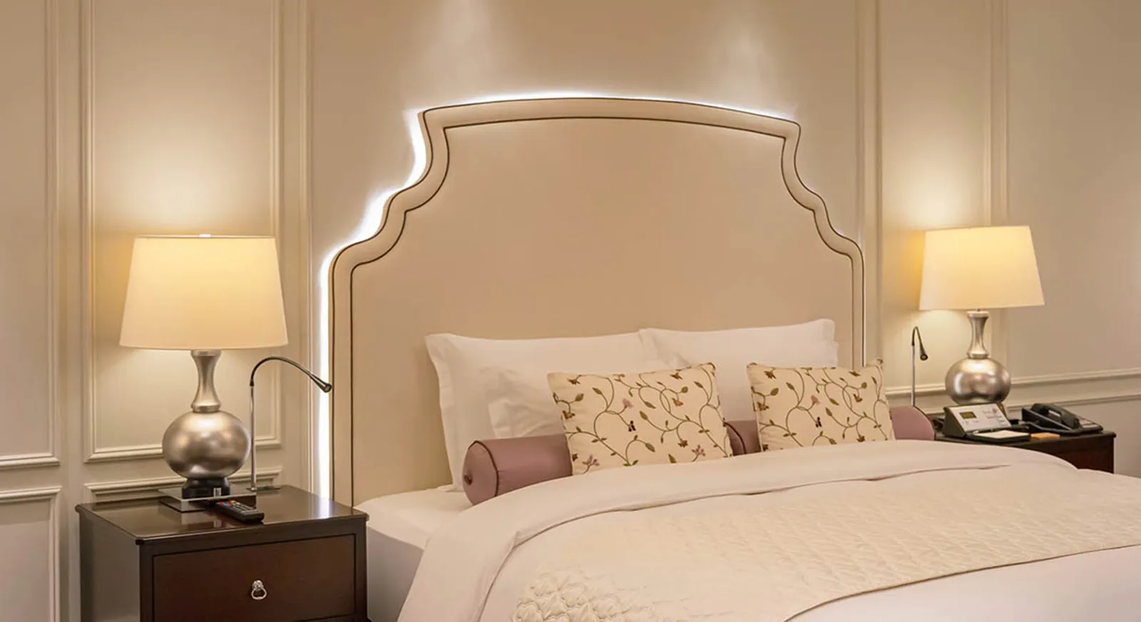 An Ambassador Suite bed, with its shaped headboard lit along the edge and 2 silver lamps.