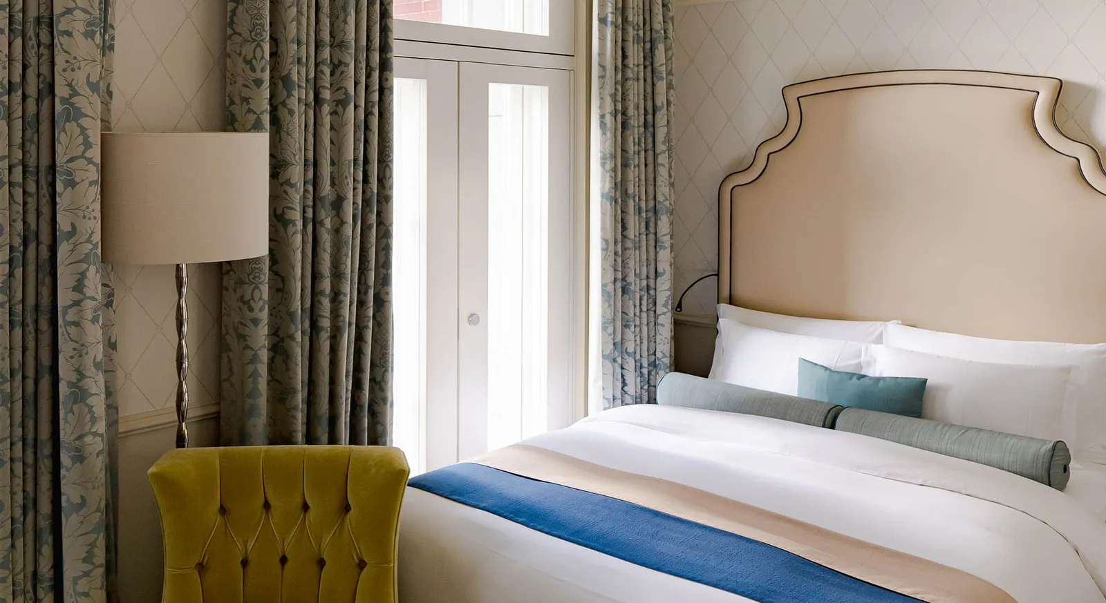 A City View room with the bed, a yellow armchair, a floor lamp and blue patterned curtains at the French window.