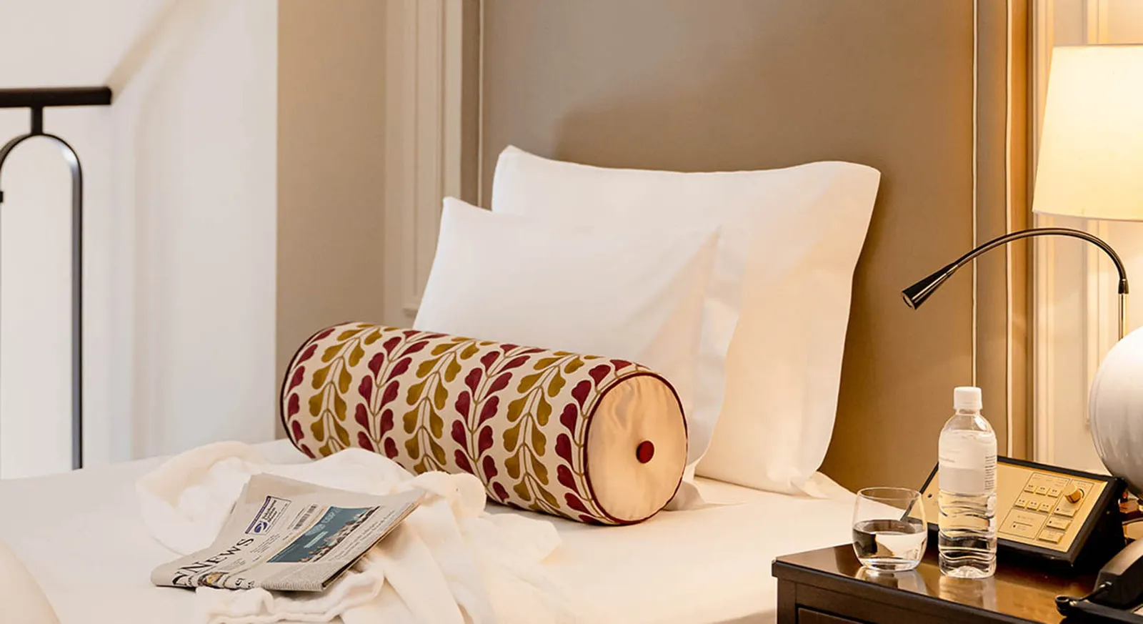 A Maisonette bed, with its red and yellow patterned bolster, a newspaper and water on the bedside table.