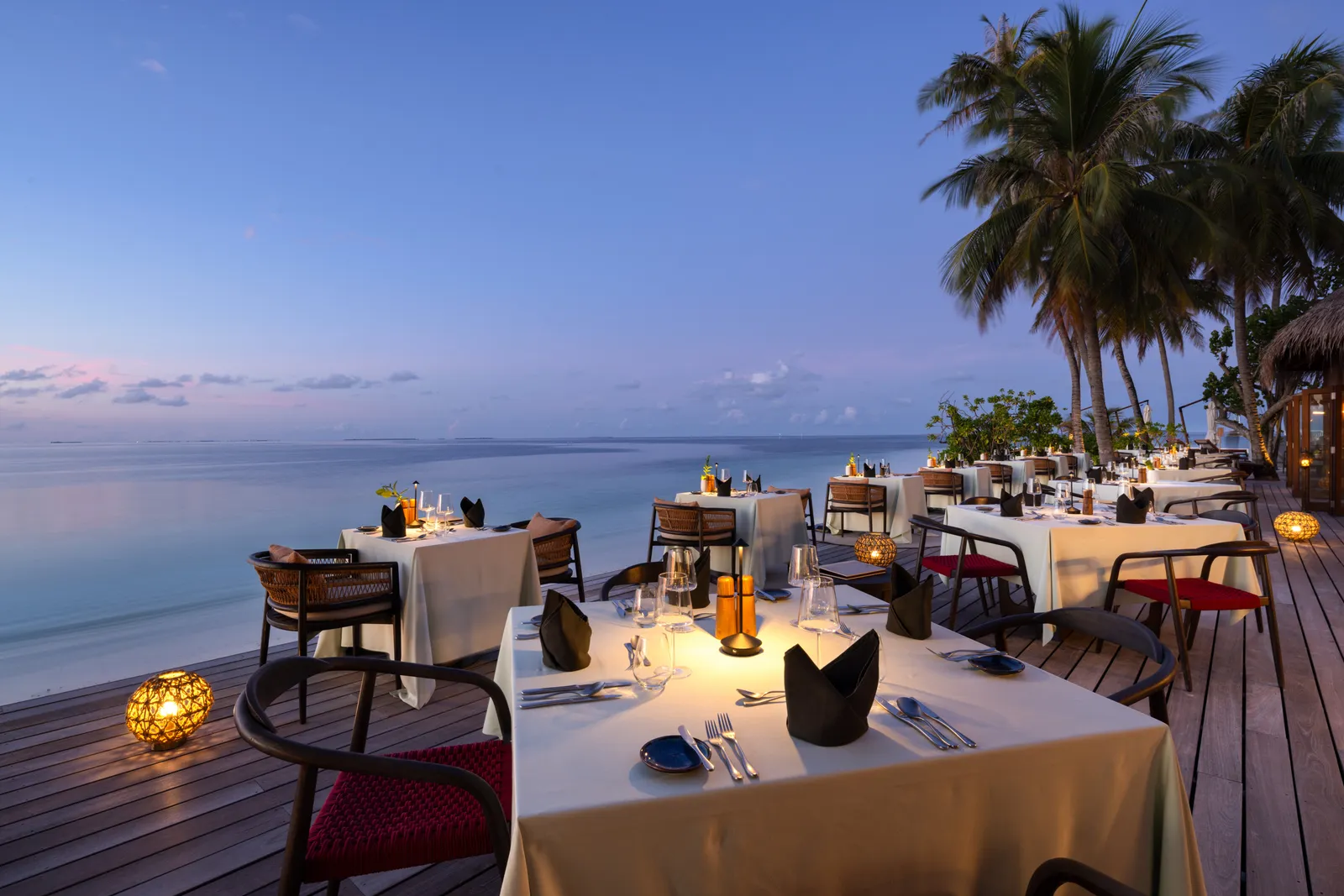 Dhonveli’s deck by the sea at dusk, tables laid, lanterns lit and palms behind.