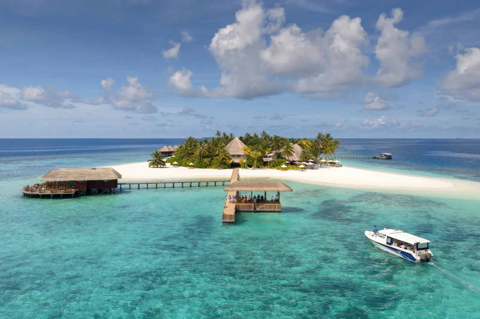Mirihi from above: the palm-covered island and its tongue of sand, the jetties, a restaurant over the water and a speedboat in the lagoon.