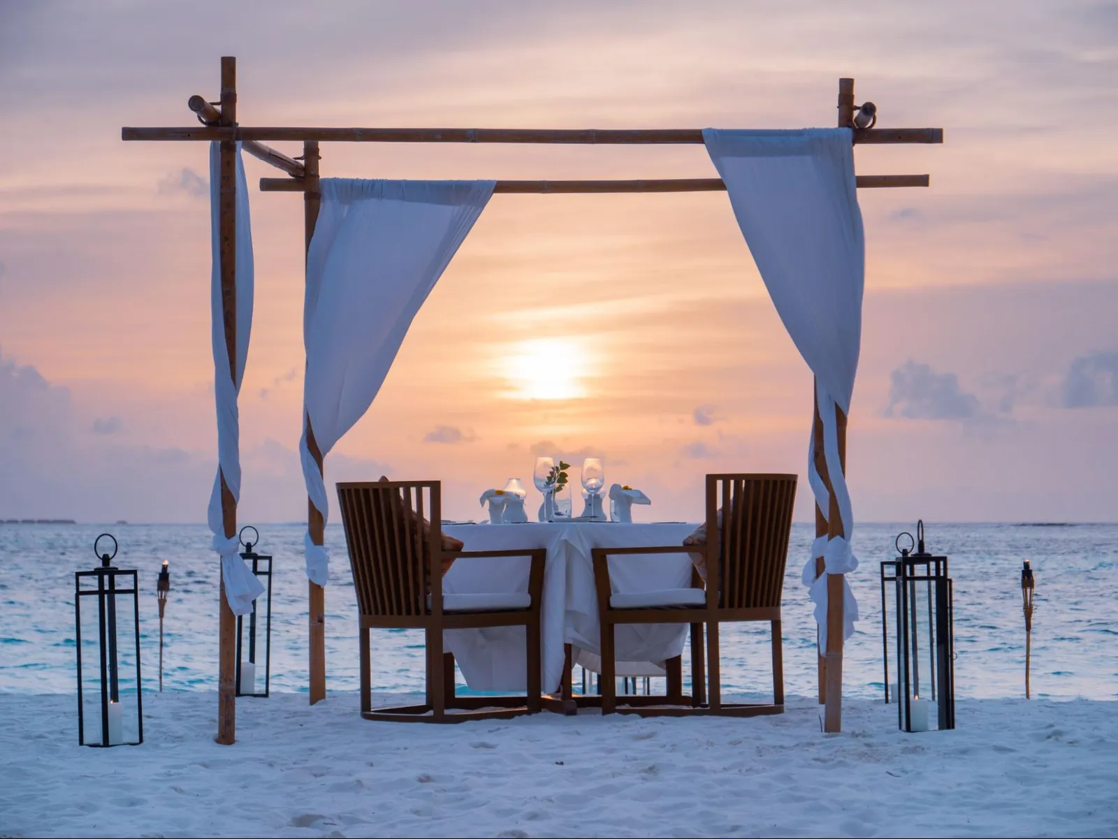 A table for 2 on a sandbank at sunset, under a frame hung with white curtains, with lanterns.
