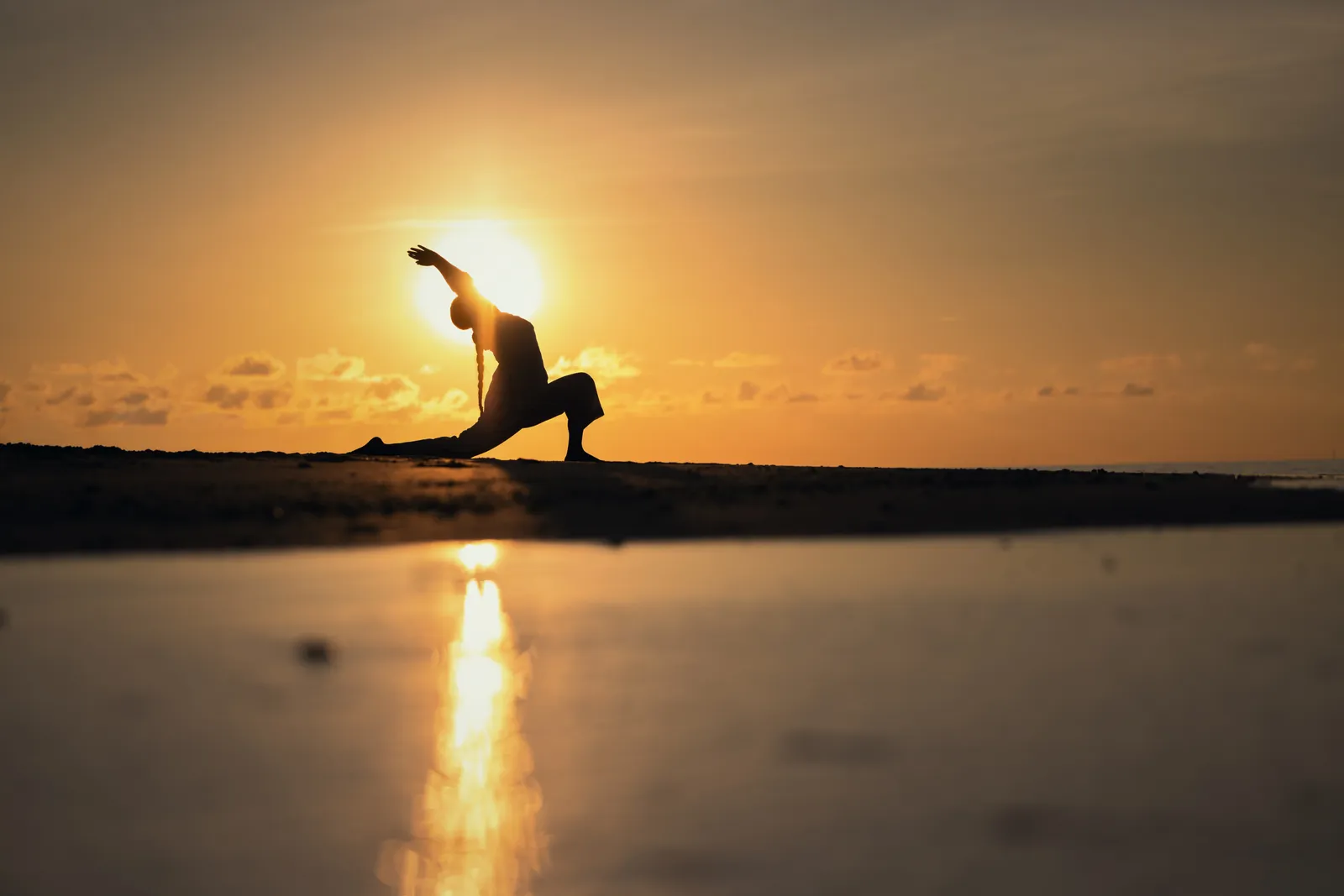 A woman in a yoga pose on the sand, silhouetted against the sun setting over the sea.