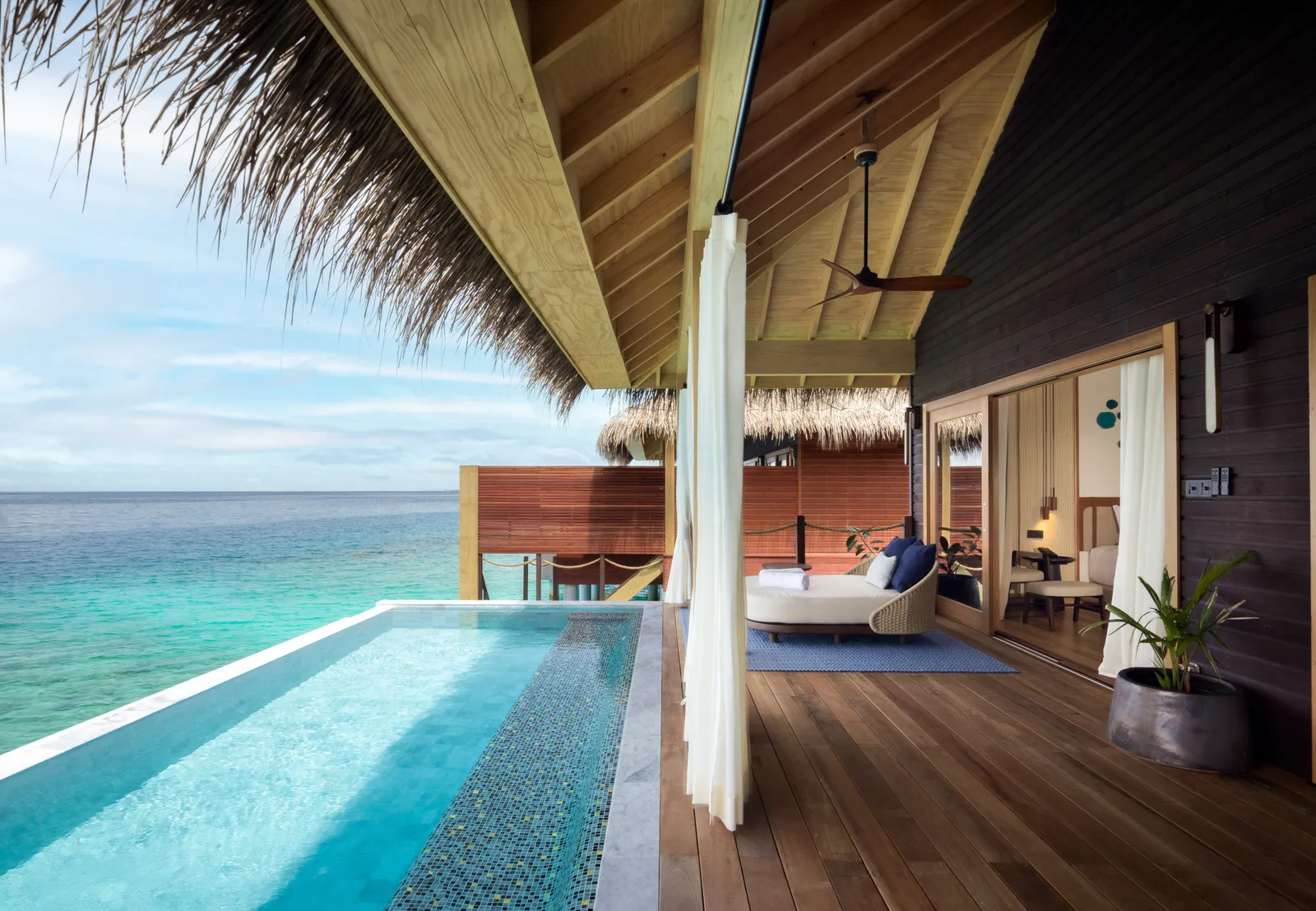 A water villa’s deck with its infinity pool, white curtains, an outdoor sofa and the sea to the horizon.