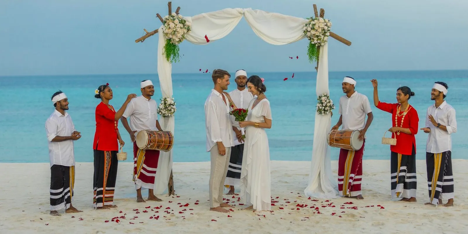 A ceremony on the beach: the couple under a flower arch, flanked by bodu beru drummers in red and white.