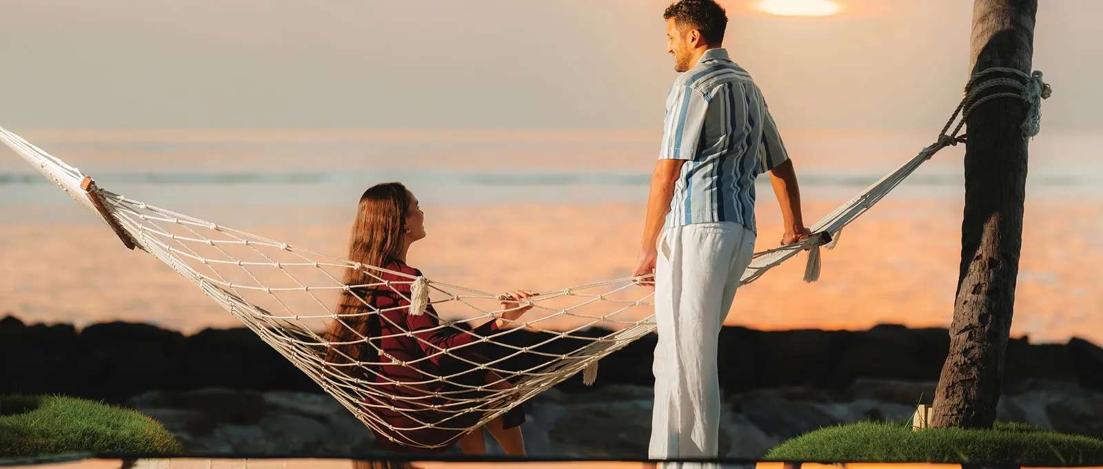 A couple at sunset by the lagoon, she lying in a rope hammock and he standing beside it.
