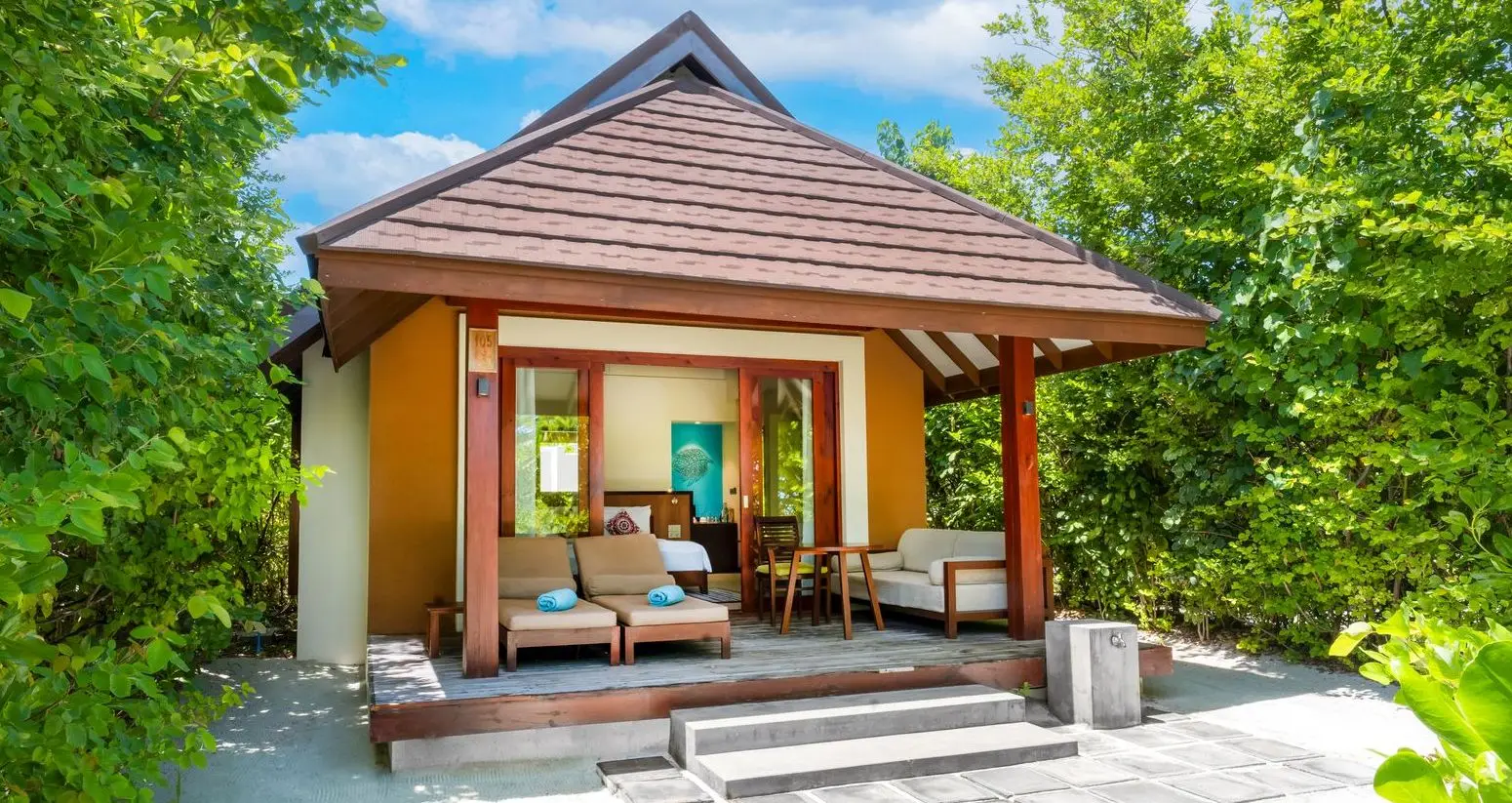 A Beach Villa among the greenery, with its timber veranda, the sofa and 2 loungers in front.