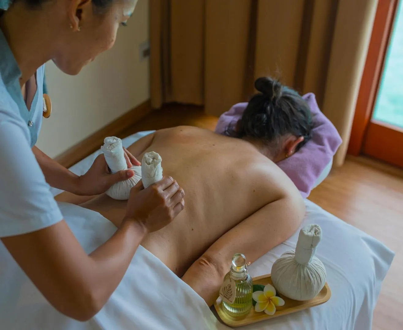 A therapist working warm sand pouches over a guest’s back in the Veli Bon’dli ritual, in an ELE|NA Ayur room.