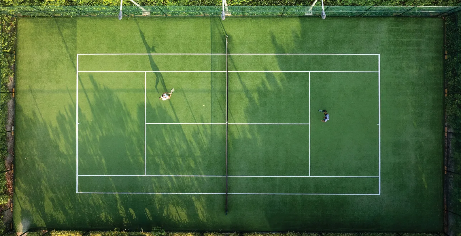 The tennis court from above, green, with 2 players and the shadows of palms along its edge.