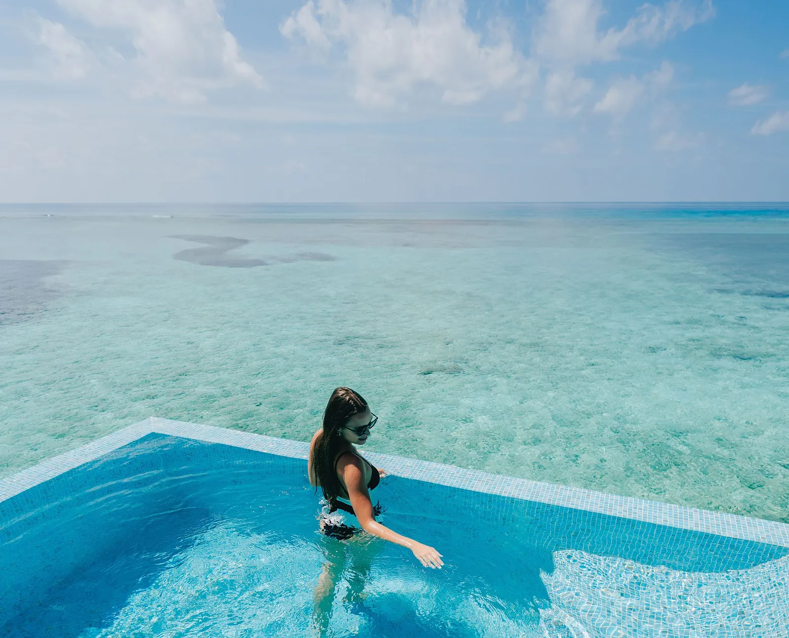 A guest wading through a water villa’s infinity pool, with the turquoise lagoon all around.