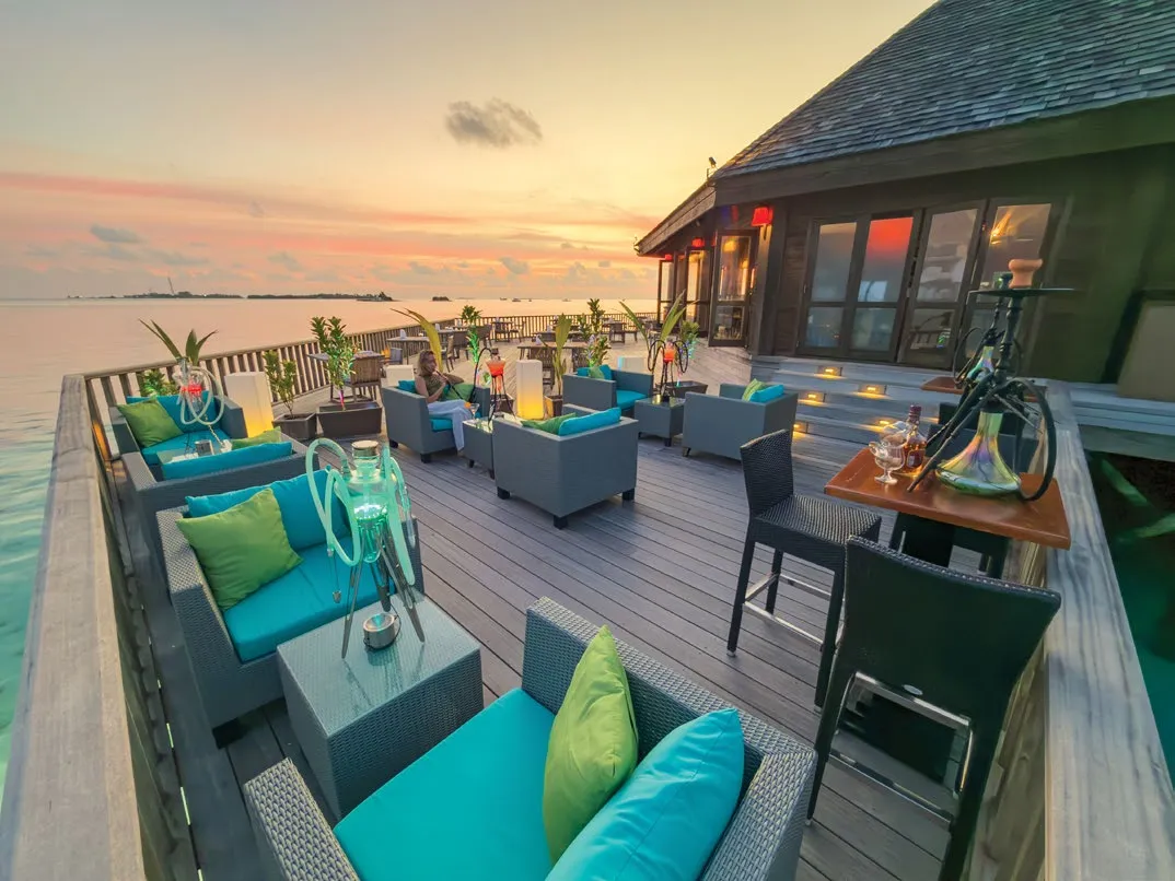 A bar deck over the water at dusk, with sofas and bright cushions.