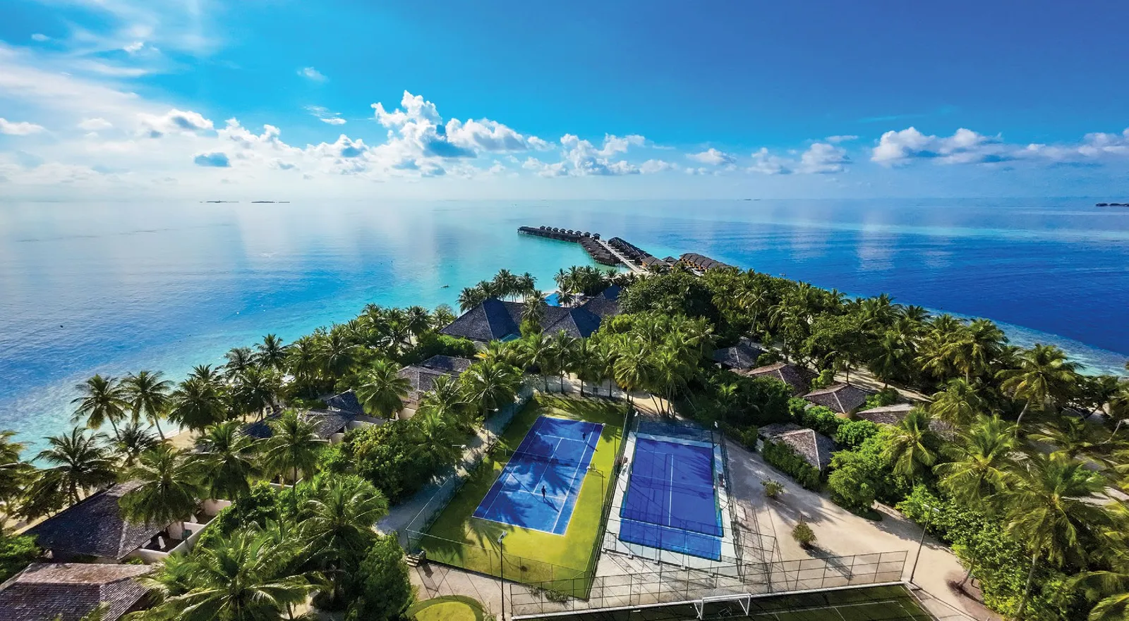 The island from above, with the tennis and padel courts among the palms and the jetty running out to sea.