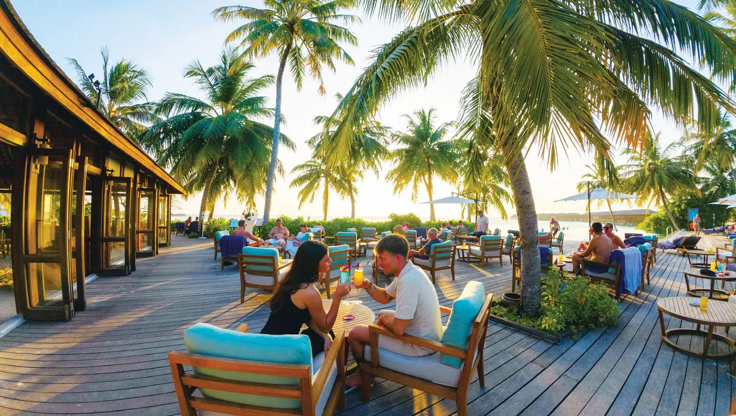 A couple raise their glasses on a timber deck among the palms, with other guests at the tables in the late afternoon light.