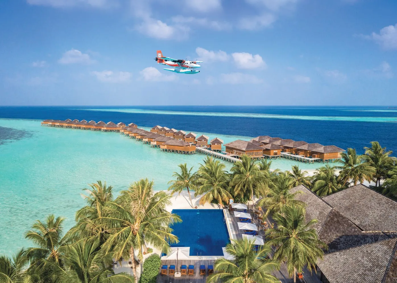 A seaplane over the lagoon, the water villas in a line and a blue pool among the palms.