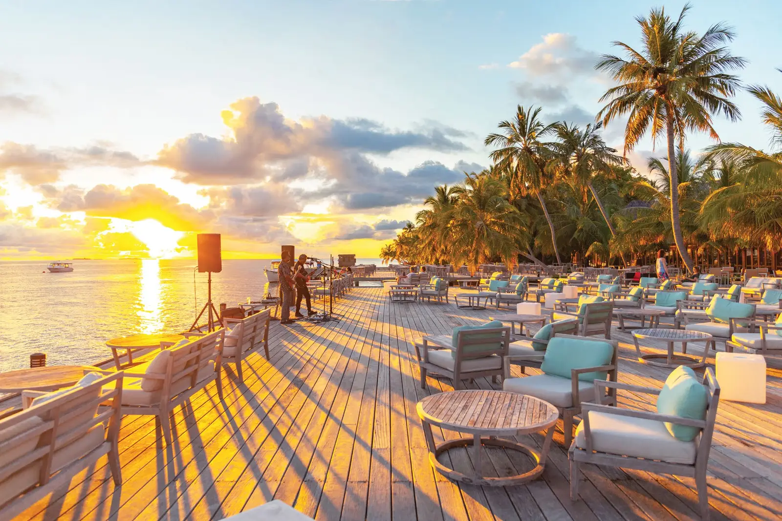 A timber deck over the sea at sunset, with sofas, palms and the sun low on the horizon.