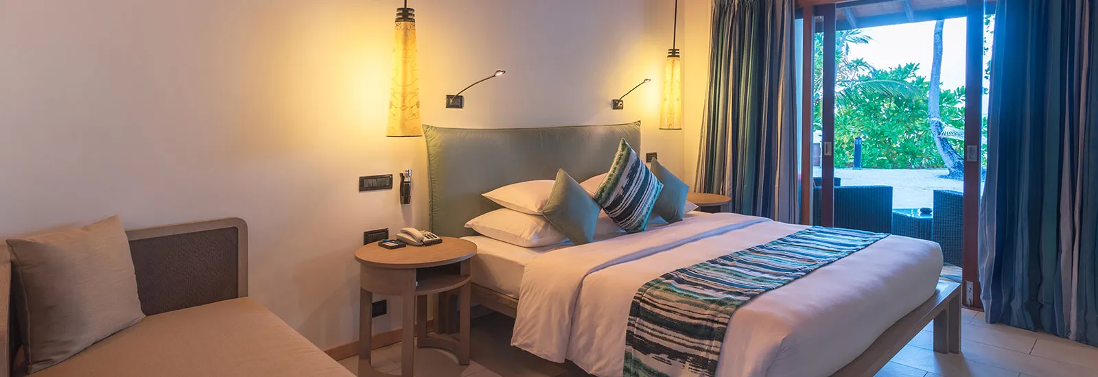 A Beachfront Villa: the king bed between 2 lamps, the sofa and the glass door open to the green by the beach.