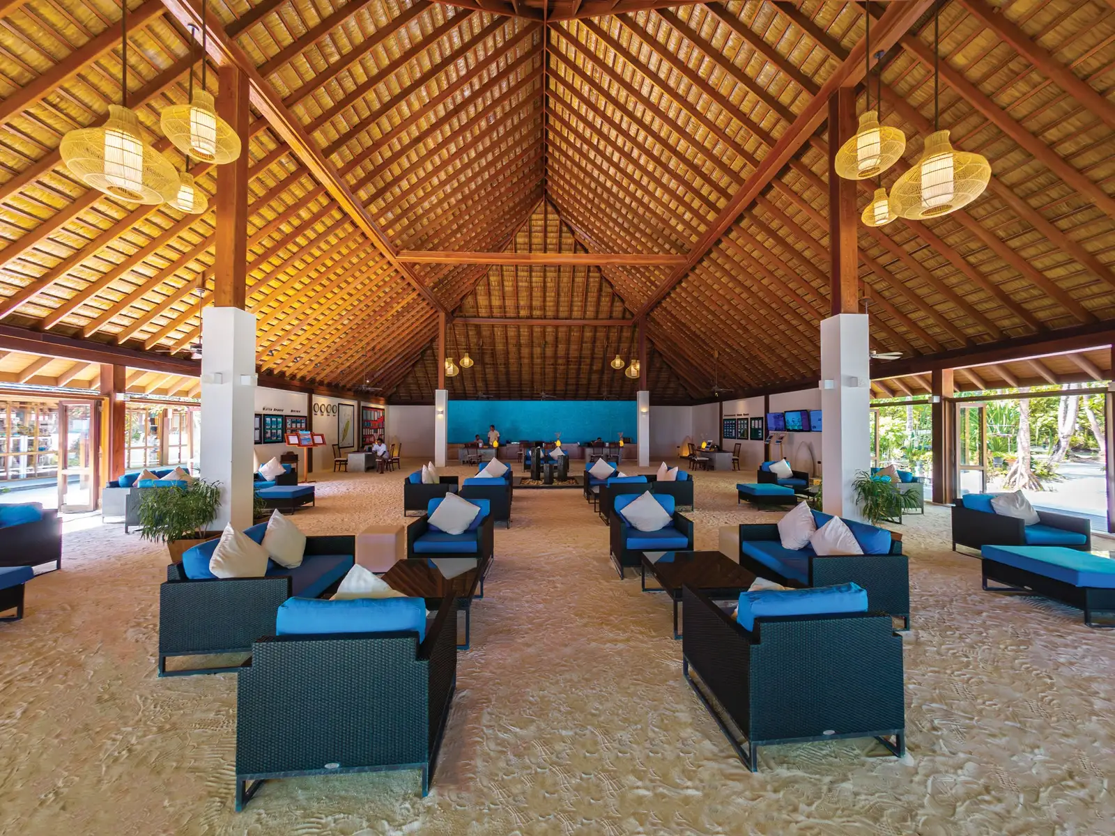 An open lounge under a high timber roof, with blue sofas on a sand floor.