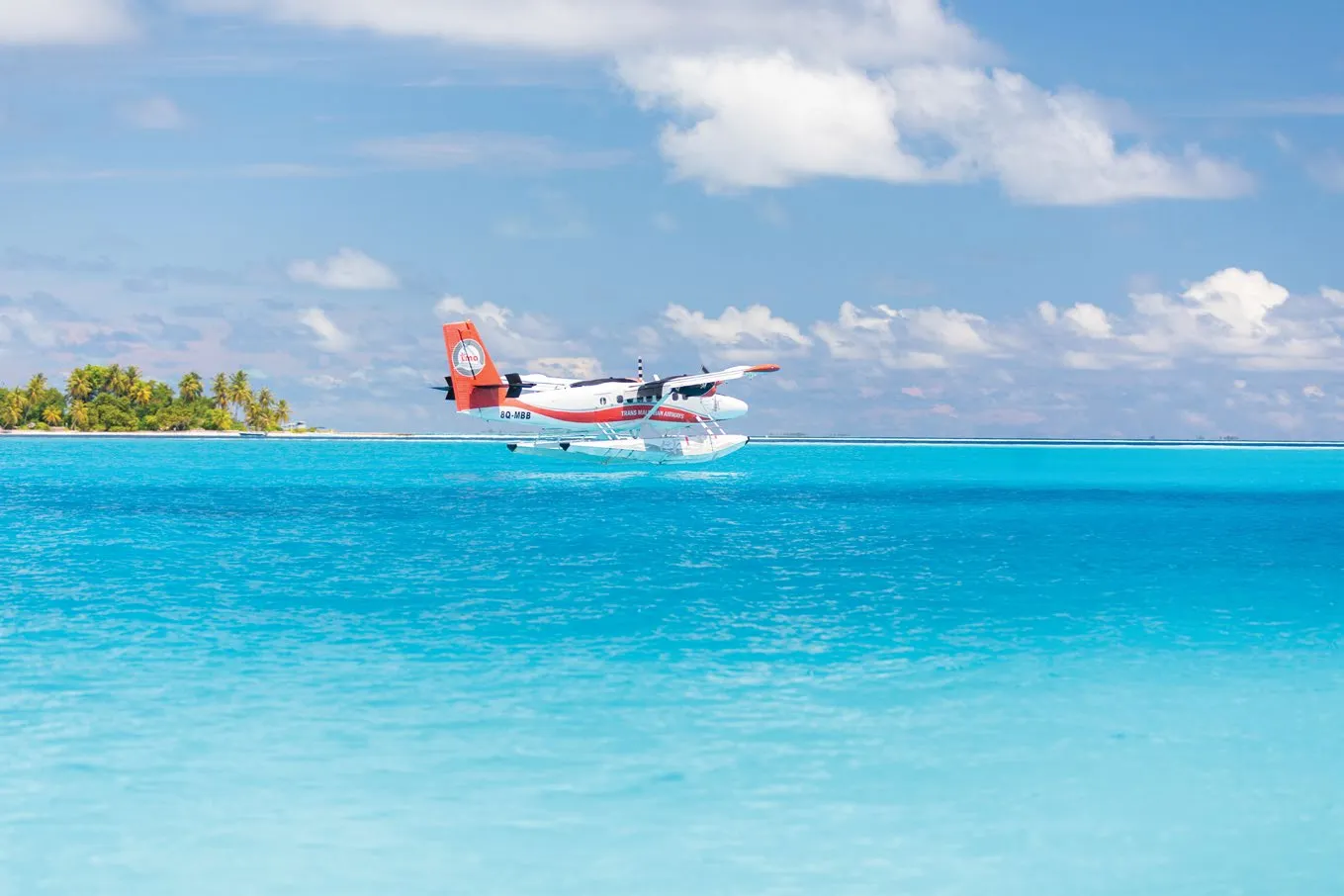 A seaplane settles on the turquoise lagoon, with a palm island behind.