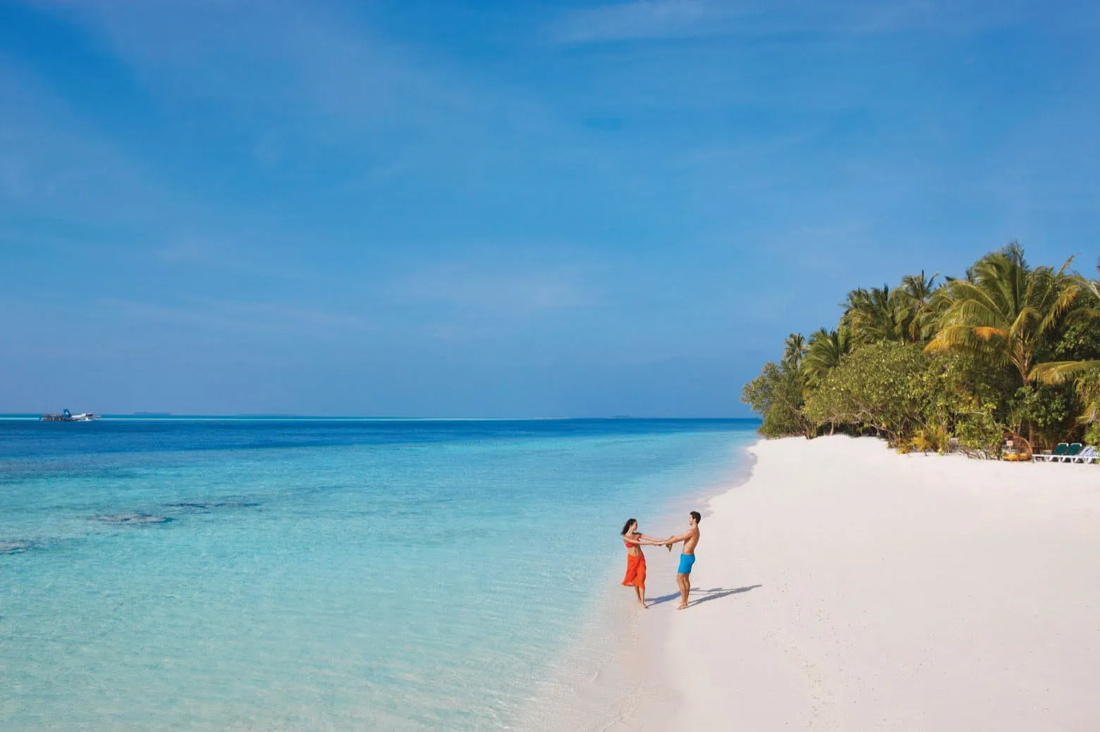 A couple holding hands on the white beach, between the turquoise water and the palms.