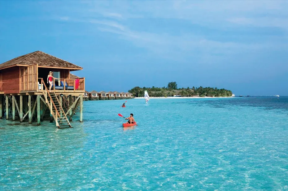 2 people kayaking in the lagoon in front of the water villas, with a sail in the distance.