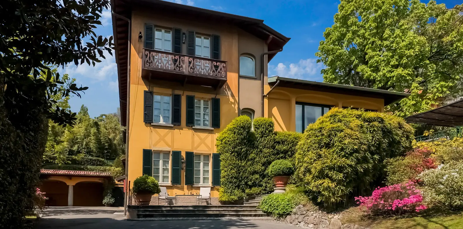 Villa Garrovo’s yellow facade with green shutters and flowering shrubs