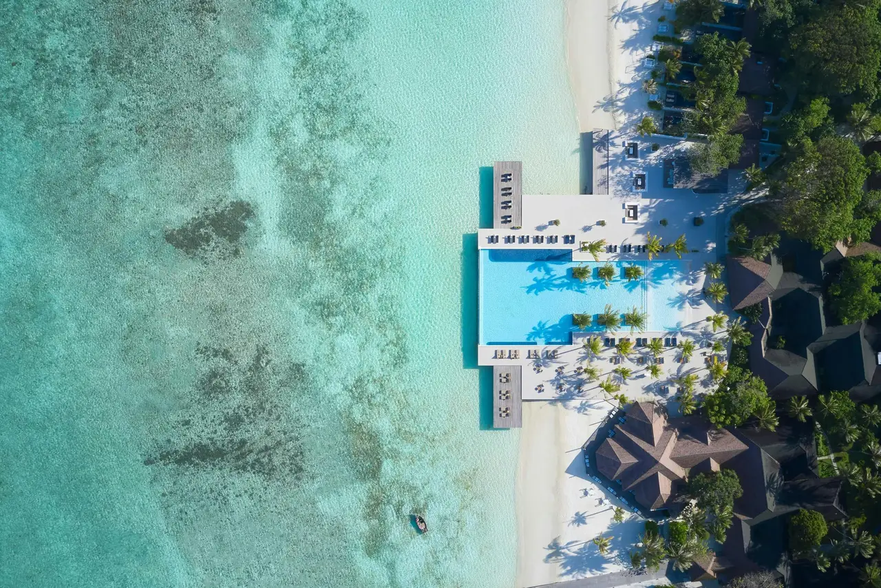 The main pool from above, between the white beach, the loungers and the reef darkening the lagoon.