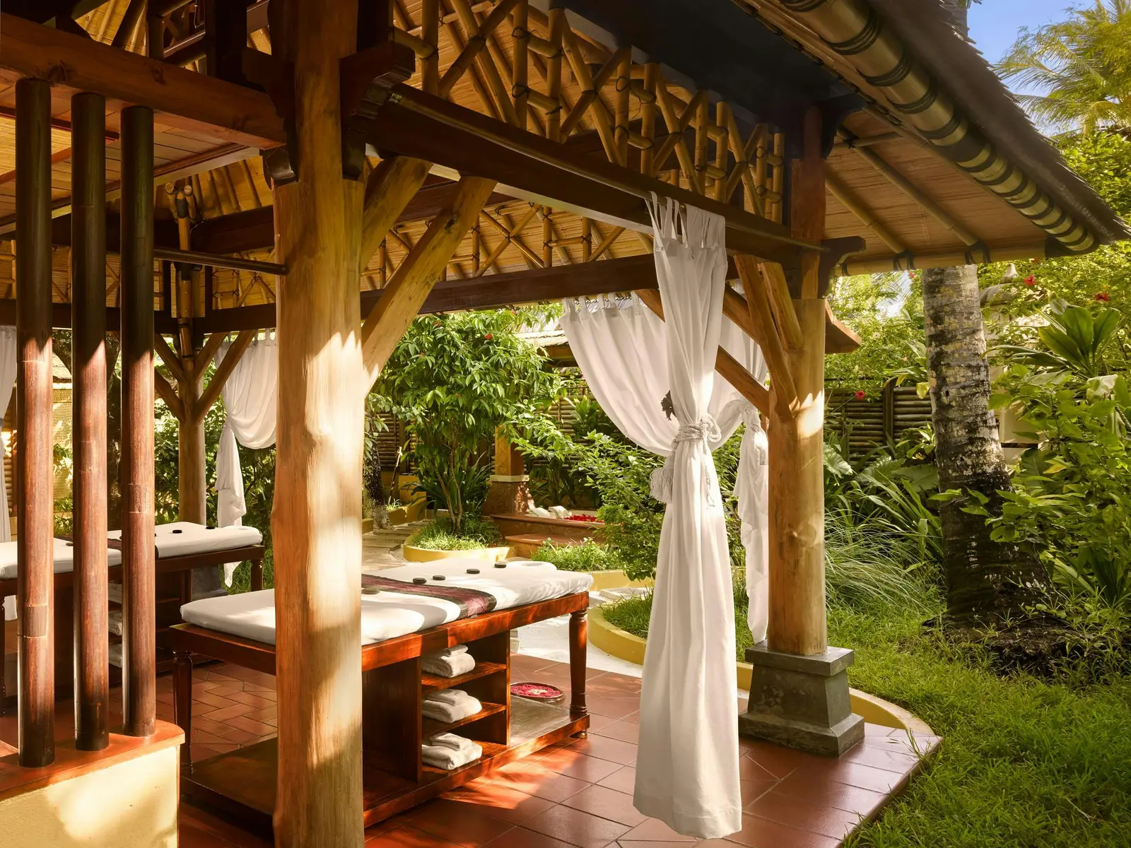 Open-air garden pavilion with 2 treatment beds, curtains, towel shelves and foliage.