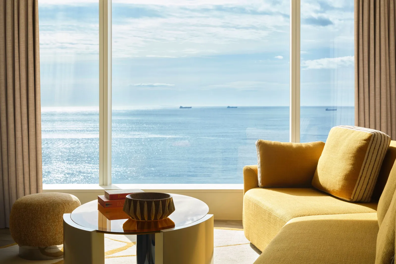 A yellow armchair and a side table at a window on the sea and ships at anchor