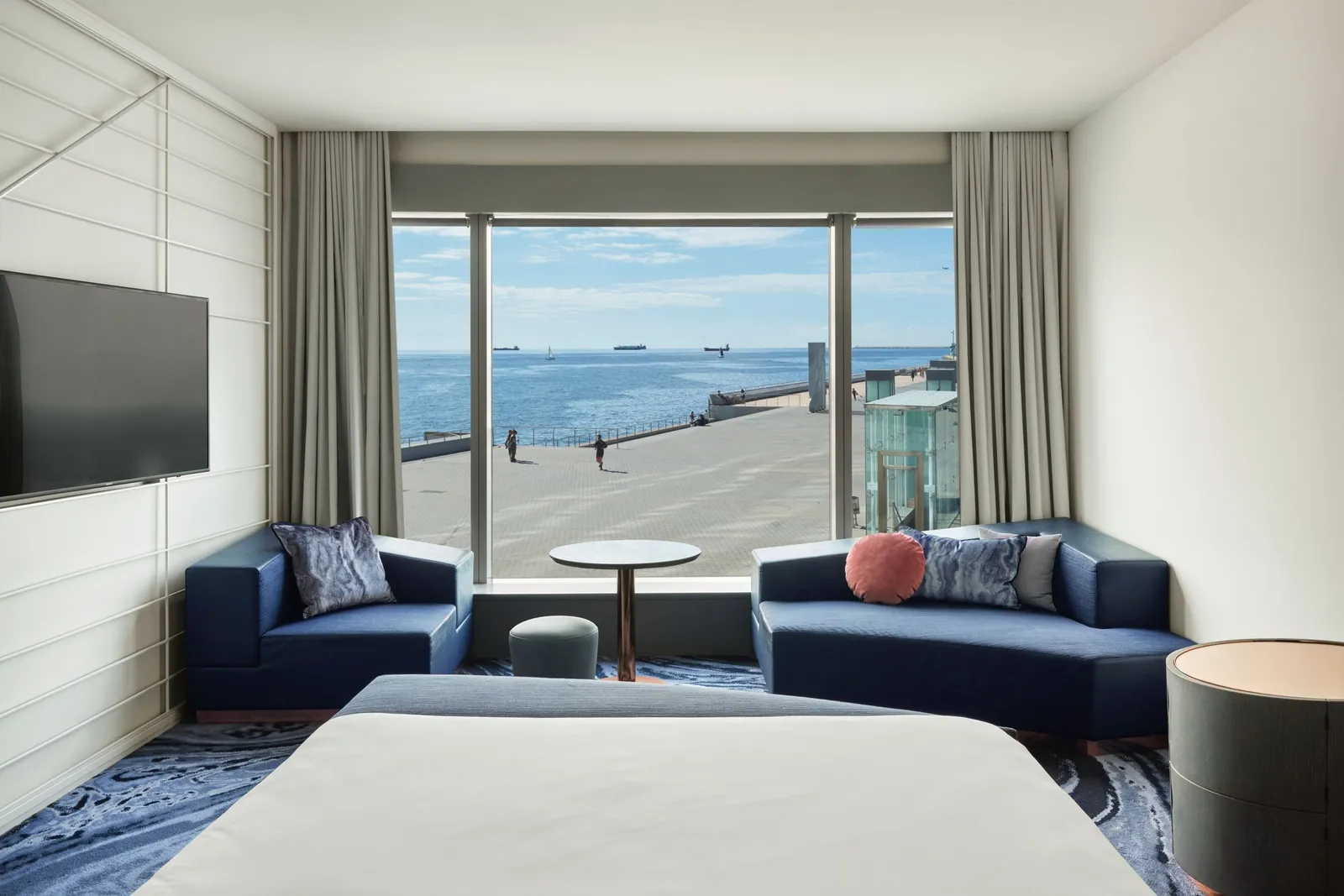 A room with a white bed, 2 small navy armchairs and a window onto the quay and the sea