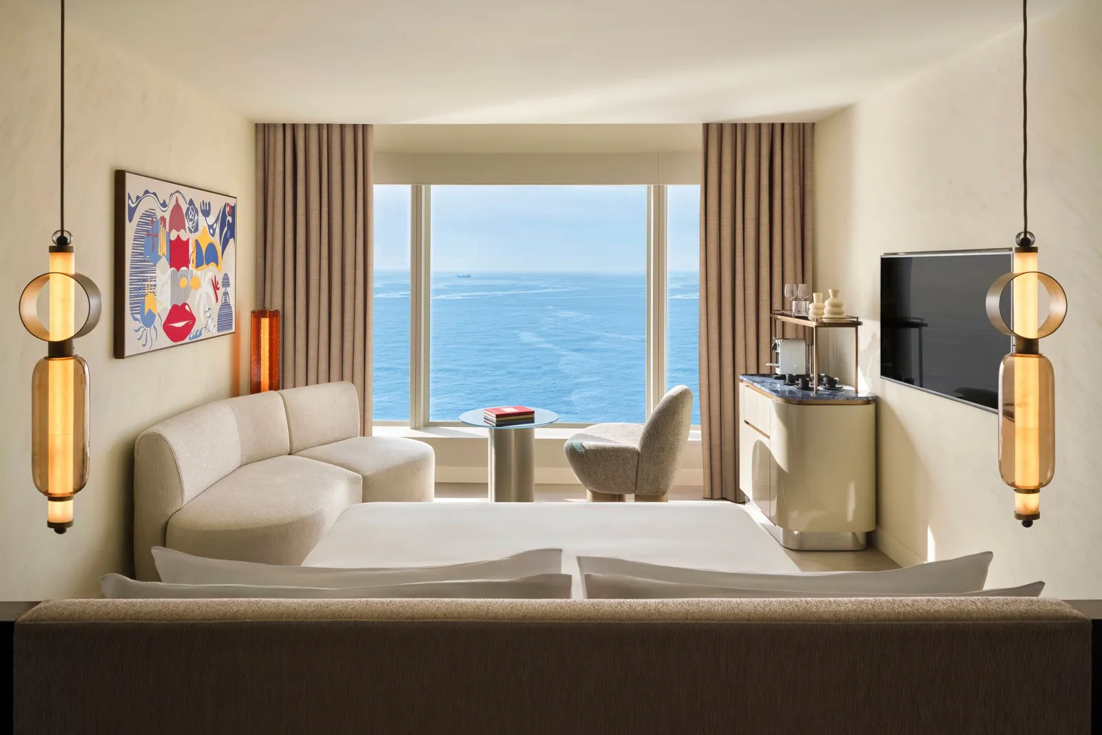 A room with a white sofa, a bright painting and the sea at the end of the window