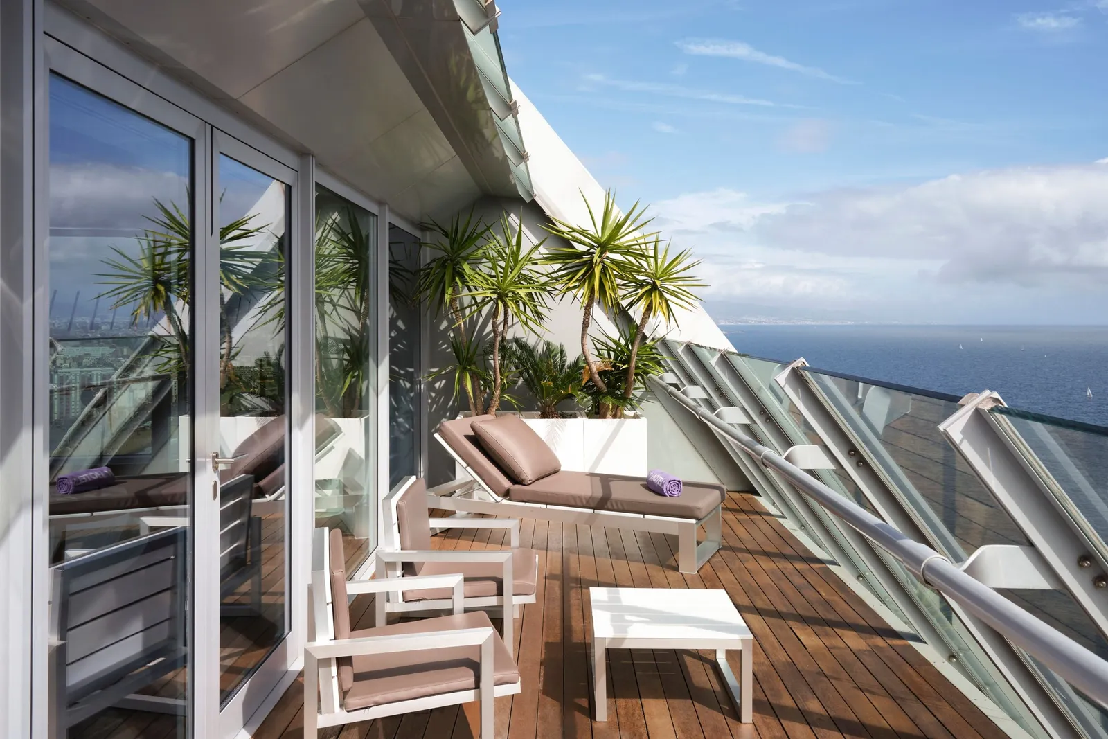 A timber terrace with a lounger and chairs, the tower’s sail above and the sea beyond