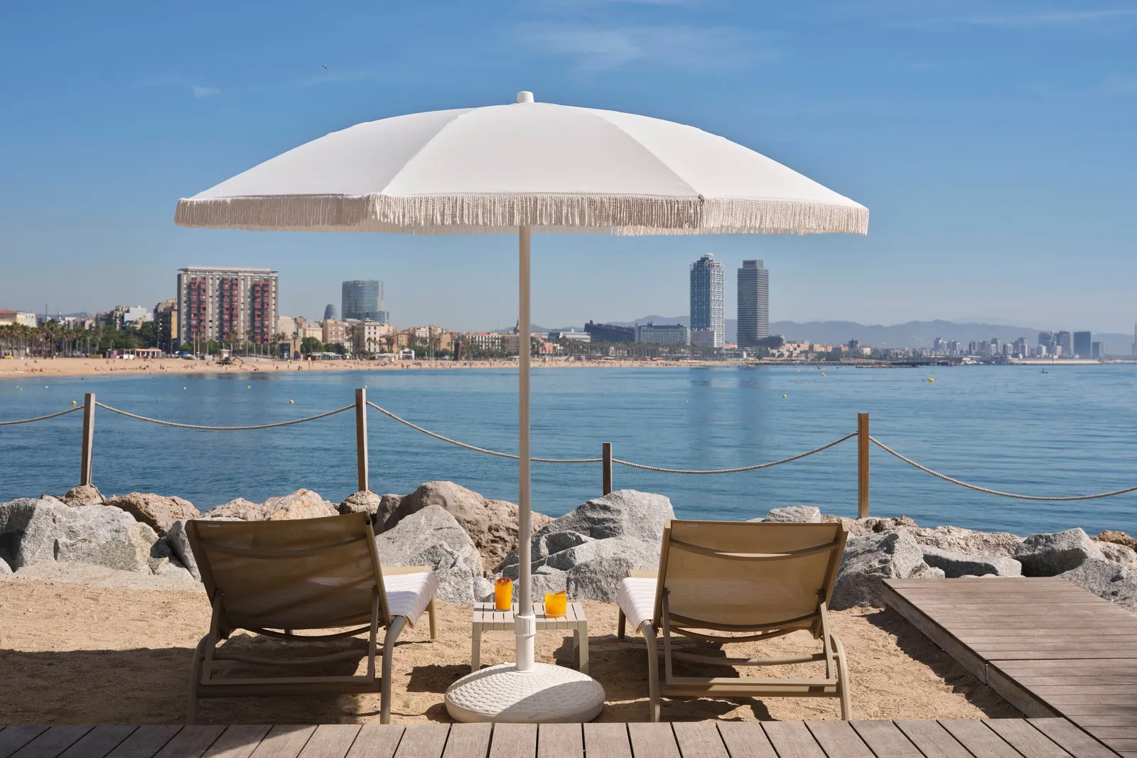 A fringed parasol and 2 loungers on SALT’s sand, with Barceloneta opposite