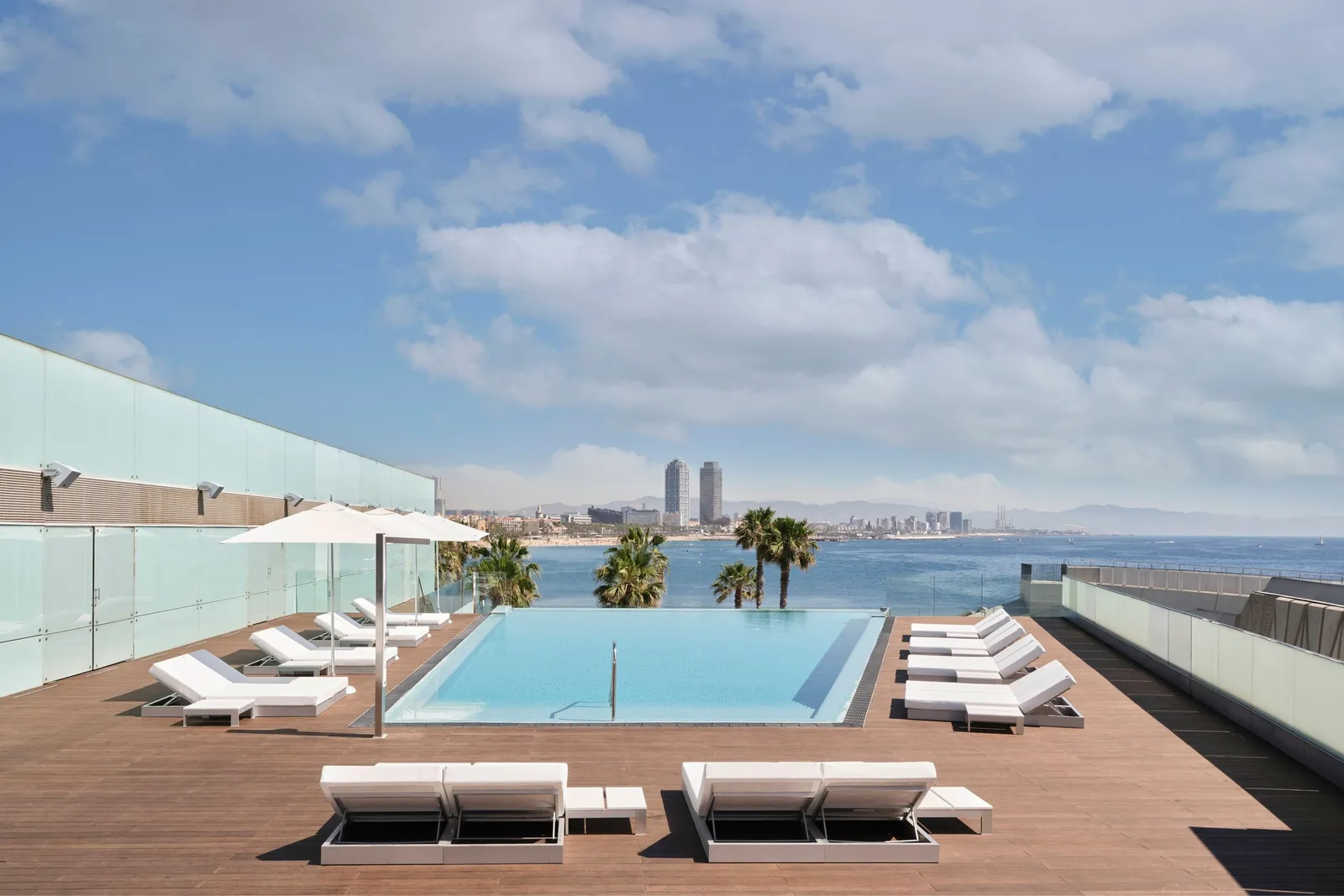 The SUN Deck infinity pool over the sea, white loungers and the 2 Port Olímpic towers in the distance