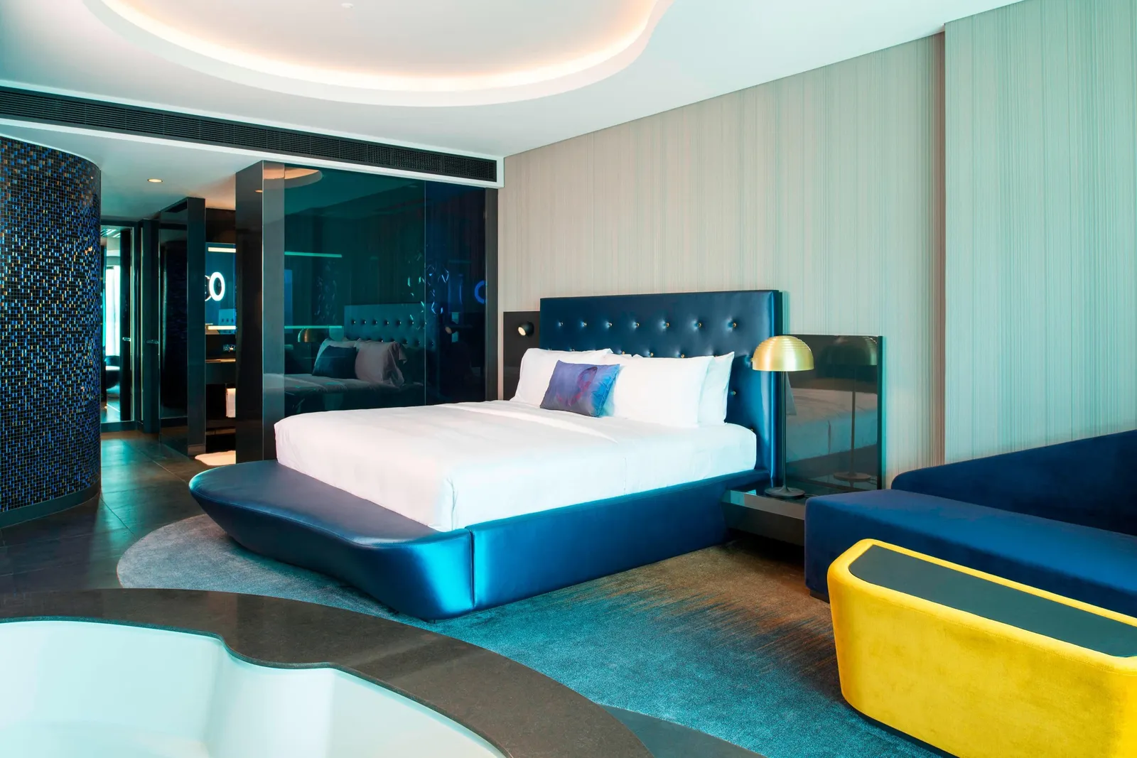 The W Suite bedroom with the bed, a yellow sofa and the bath behind glass
