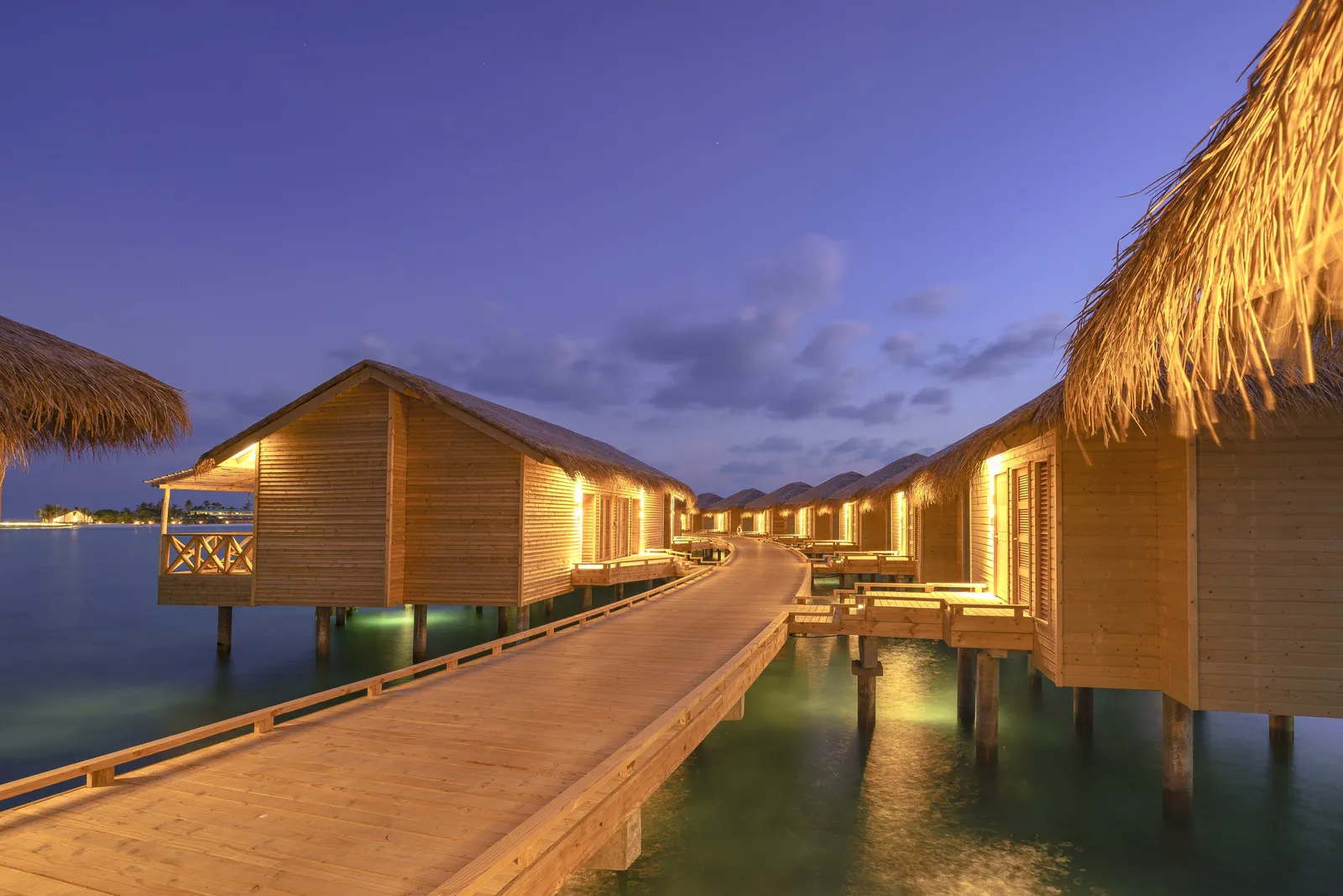 The water-villa walkway at dusk, lights on beneath the thatched roofs